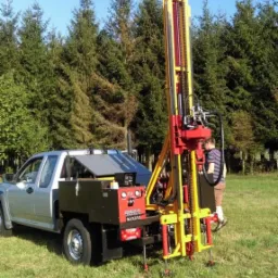 Wiertnica zamontowana na pickupie podczas pracy na zielonej łące, w tle widoczne drzewa iglaste i błękitne niebo.