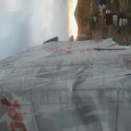 Szkielet konstrukcji budynku pokryty białą siatką ochronną z czerwonym nadrukiem, w tle widoczne budynki i zachodzące słońce.