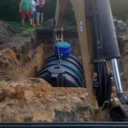 Żółta koparka CAT opuszcza czarny zbiornik z niebieską pokrywą do wykopu w ziemi, w tle obserwują trzy osoby.