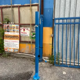 Niebieski słupek z zamontowanymi elementami hydraulicznymi na tle blaszanej ściany i plakatu reklamowego Ceramizer, potencjalnie element systemu kontroli dostępu.