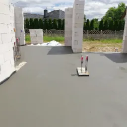 Świeżo wylana, gładka posadzka betonowa wewnątrz budynku w trakcie budowy. Widoczne bloki z betonu komórkowego tworzące fragment ściany oraz pionowe pręty z zaznaczonym poziomem wylewki.