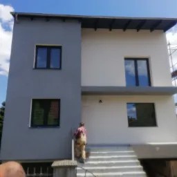 Dwukondygnacyjny dom z szarą i białą elewacją, pracownik w kombinezonie wchodzi po schodach wejściowych, widoczne czarne ramy okienne i fragment rusztowania.