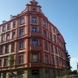 Odnowiona kamienica narożna w odcieniach pomarańczy i beżu, widok z dołu na fasadę z balkonem, antenami satelitarnymi i szyldem piekarni na parterze.