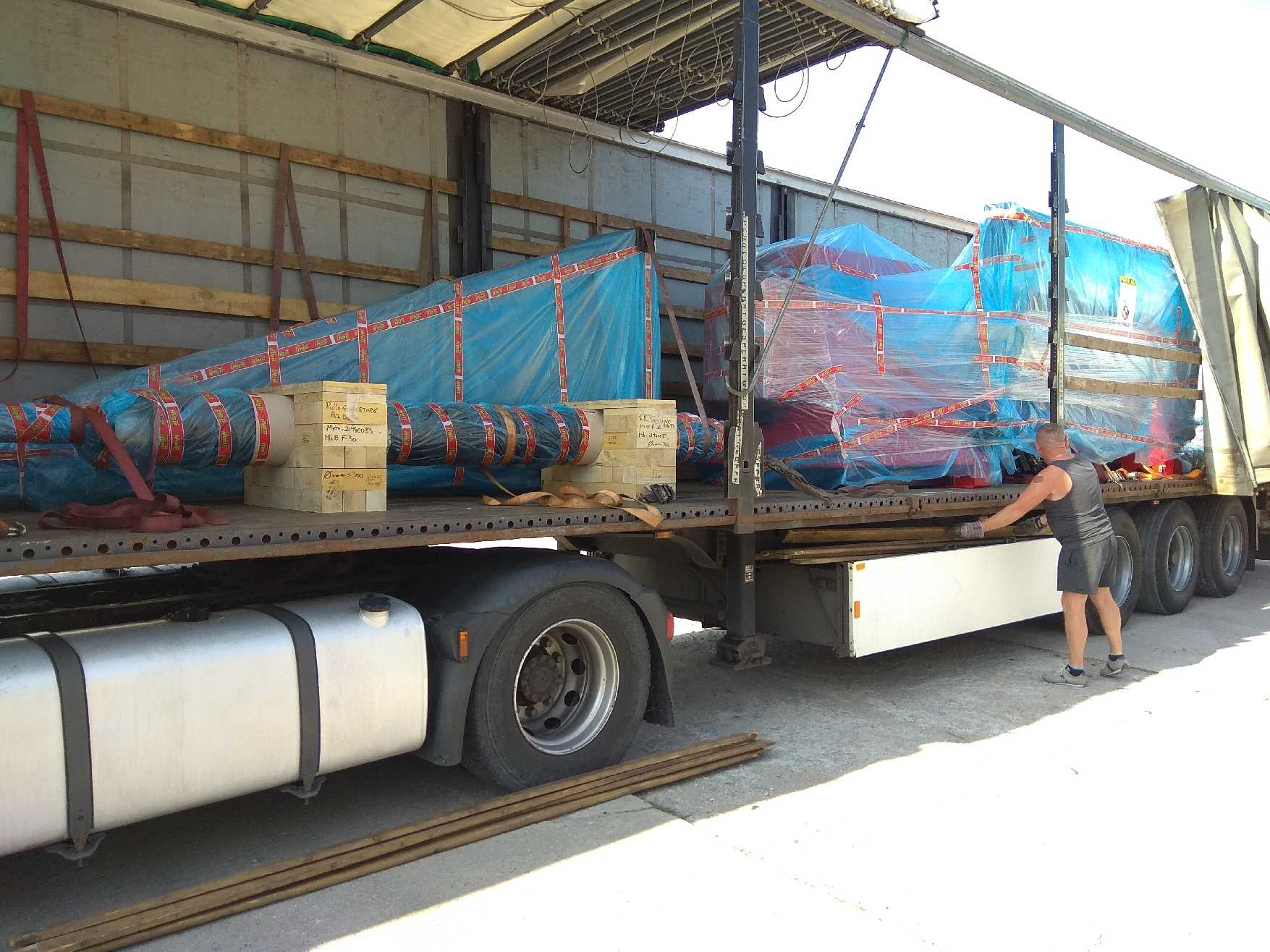 Ciężarówka z naczepą przewożąca zabezpieczone folią i taśmami ładunki na paletach, dodatkowo podparte drewnianymi klockami. Mężczyzna w szarym ubraniu manipuluje przy naczepie.