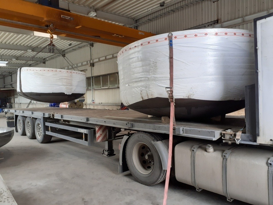 Załadunek dwóch dużych, okrągłych zbiorników zabezpieczonych białą folią na platformę transportową za pomocą suwnicy w hali przemysłowej.