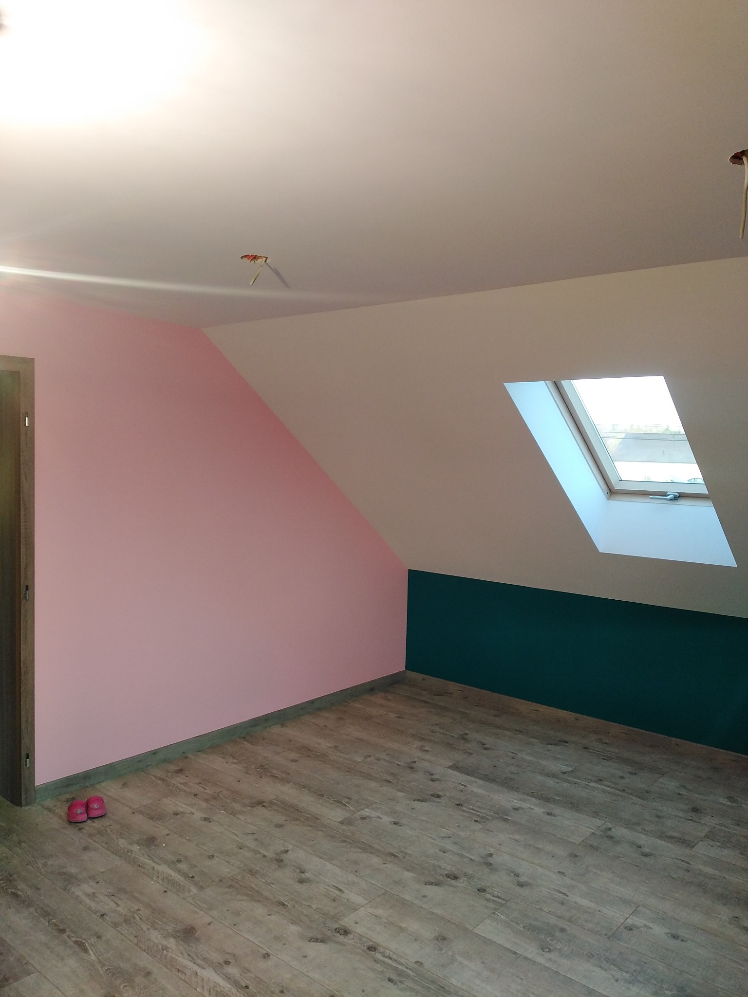 Pokój na poddaszu po malowaniu ścian na różowo i zielono, z widocznym oknem dachowym i drewnopodobną podłogą, w rogu leżą różowe kapcie.