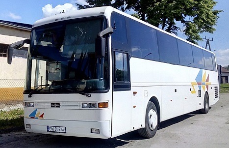 Biały autokar turystyczny z kolorowymi akcentami na boku i przodzie, zaparkowany na utwardzonym terenie przed budynkiem.