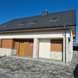 Nowoczesny dom jednorodzinny z szarą dachówką, kremową elewacją i elementami dekoracyjnymi z kamienia. Widoczne rolety zewnętrzne i kostka brukowa na podjeździe.