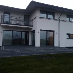 Nowoczesny, dwukondygnacyjny dom z białym tynkiem, dużymi oknami i balkonem z czarną balustradą, otoczony brukowanym tarasem i zielonym trawnikiem.