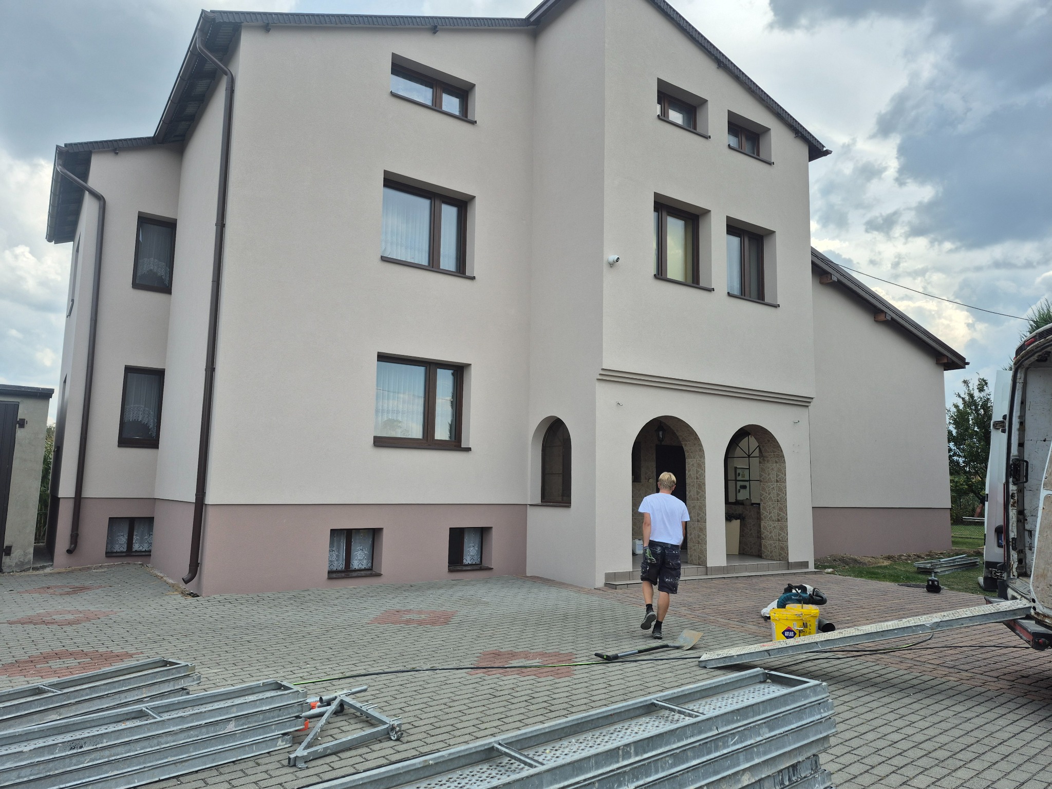 Dwukondygnacyjny dom po renowacji elewacji w odcieniach beżu i brązu, z widocznymi rusztowaniami i pracownikiem wchodzącym do budynku.