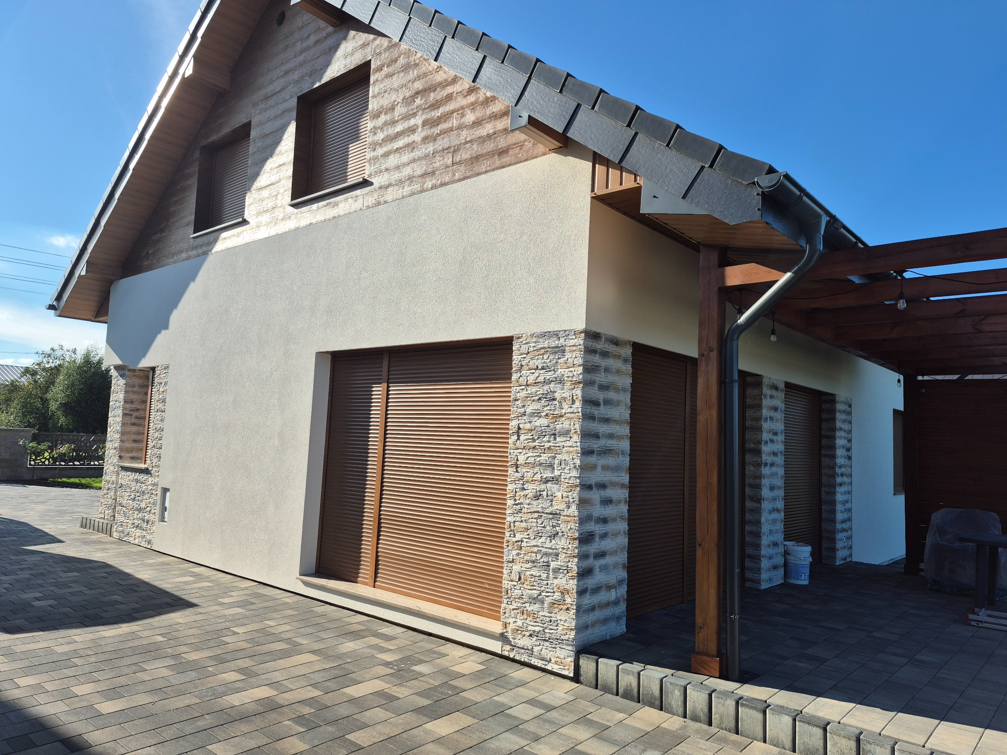 Dom jednorodzinny po remoncie elewacji: jasny tynk, okładzina z kamienia, drewniane elementy, rolety zewnętrzne. Widok z zewnątrz, kostka brukowa.
