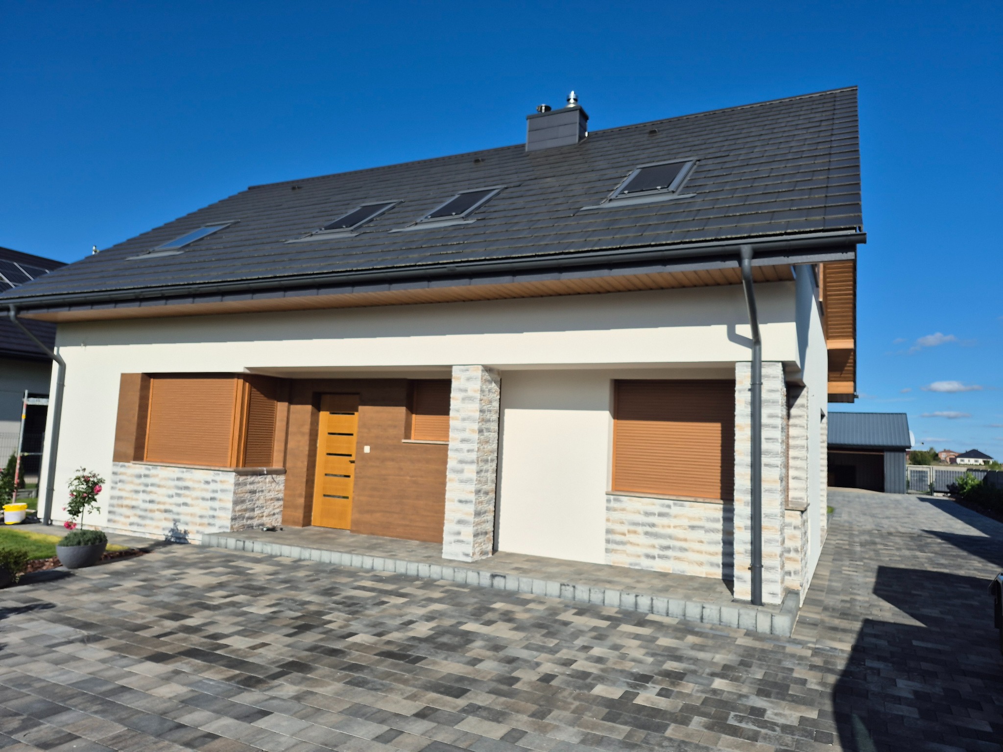 Nowoczesny dom jednorodzinny z szarą dachówką, kremową elewacją i elementami dekoracyjnymi z kamienia. Widoczne rolety zewnętrzne i kostka brukowa na podjeździe.