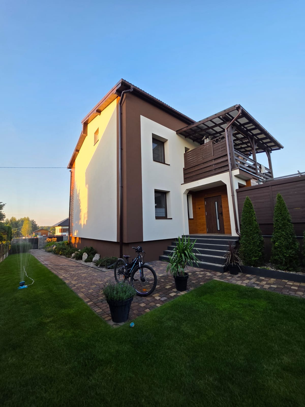 Dwukondygnacyjny dom z balkonem, elewacja w bieli i brązie, kostka brukowa, zieleń. Widok na fasadę w popołudniowym słońcu, rower oparty o donicę.