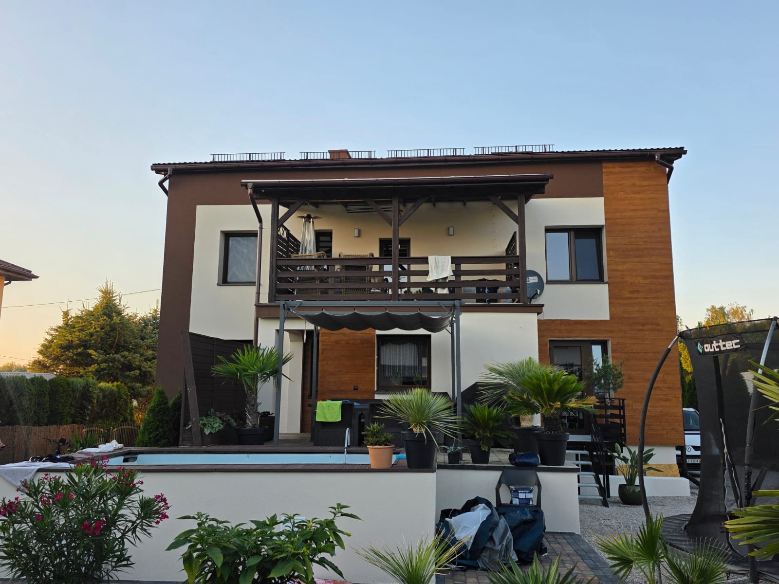Dwukondygnacyjny dom z balkonem, wykończony w tonacji beżowo-brązowej, z elementami drewnianymi. Widoczne rośliny w donicach, basen i trampolina w ogrodzie.