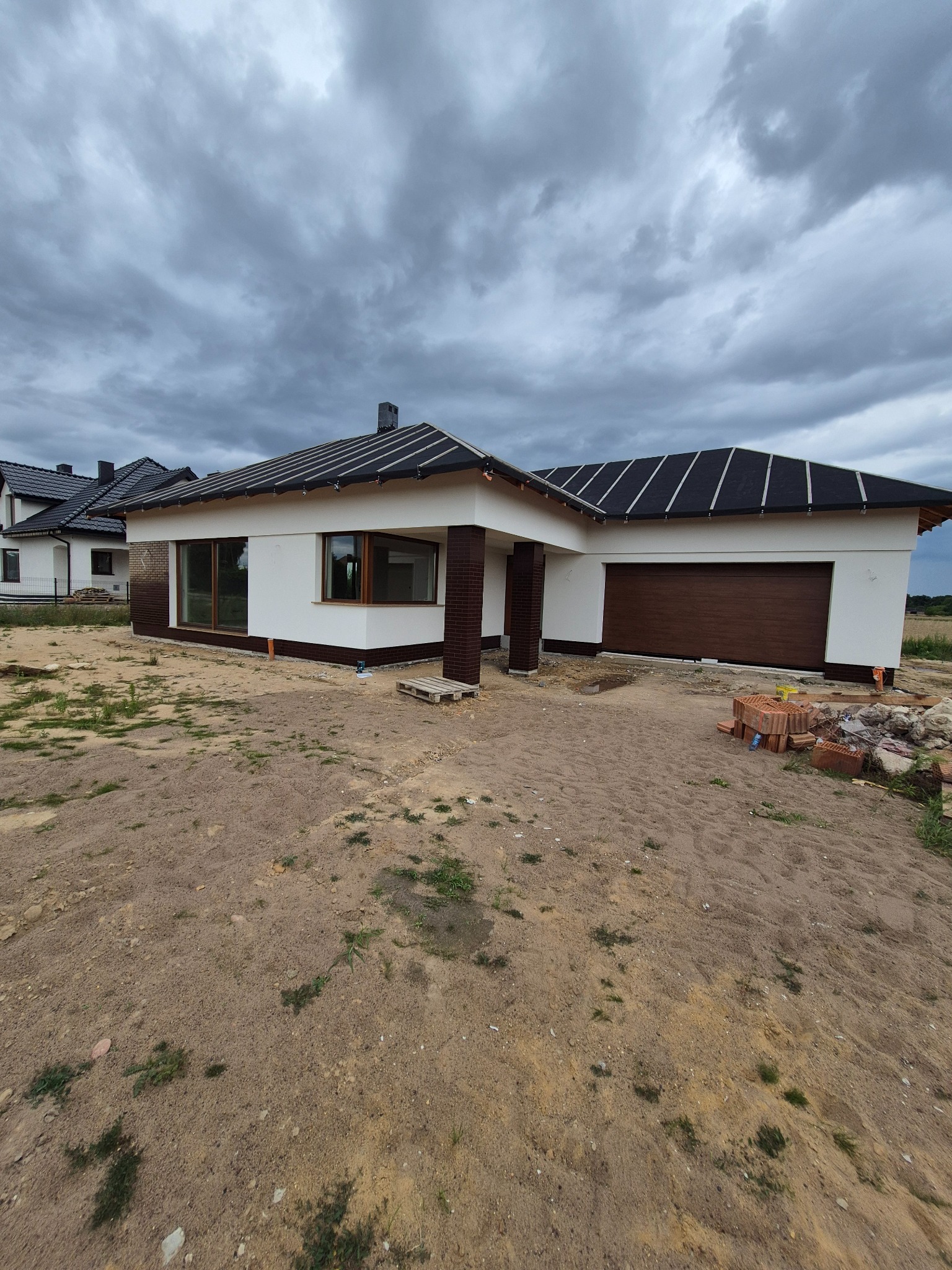 Parterowy dom jednorodzinny z brązową bramą garażową i wykończeniem elewacji imitującym cegłę, w trakcie prac wykończeniowych, na tle pochmurnego nieba.