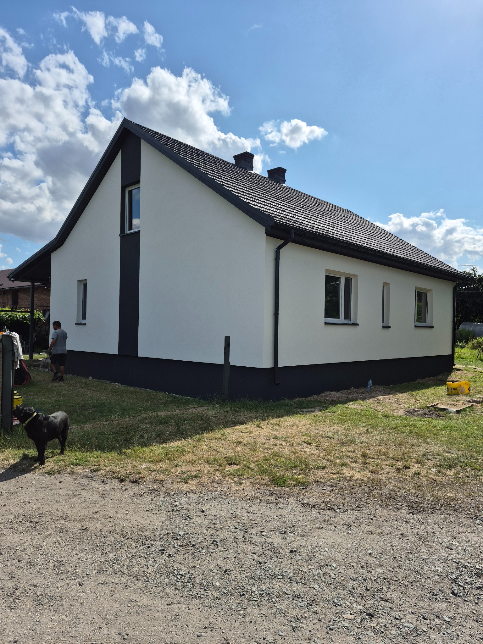 Nowoczesny dom po renowacji elewacji, z białymi ścianami i grafitowymi akcentami. Widoczny fragment ogrodu i osoba z psem w tle.