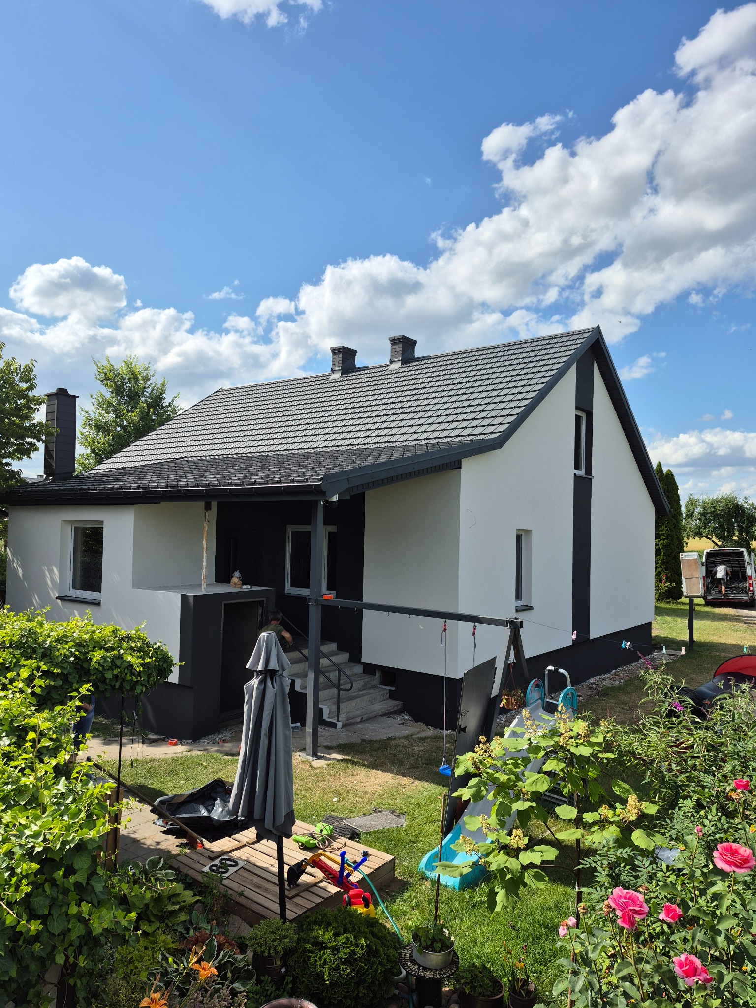 Biało-czarny dom jednorodzinny z szarą dachówką, widoczny z ogrodu pełnego zieleni i kwiatów w słoneczny dzień. Częściowo widoczny pracownik na schodach.
