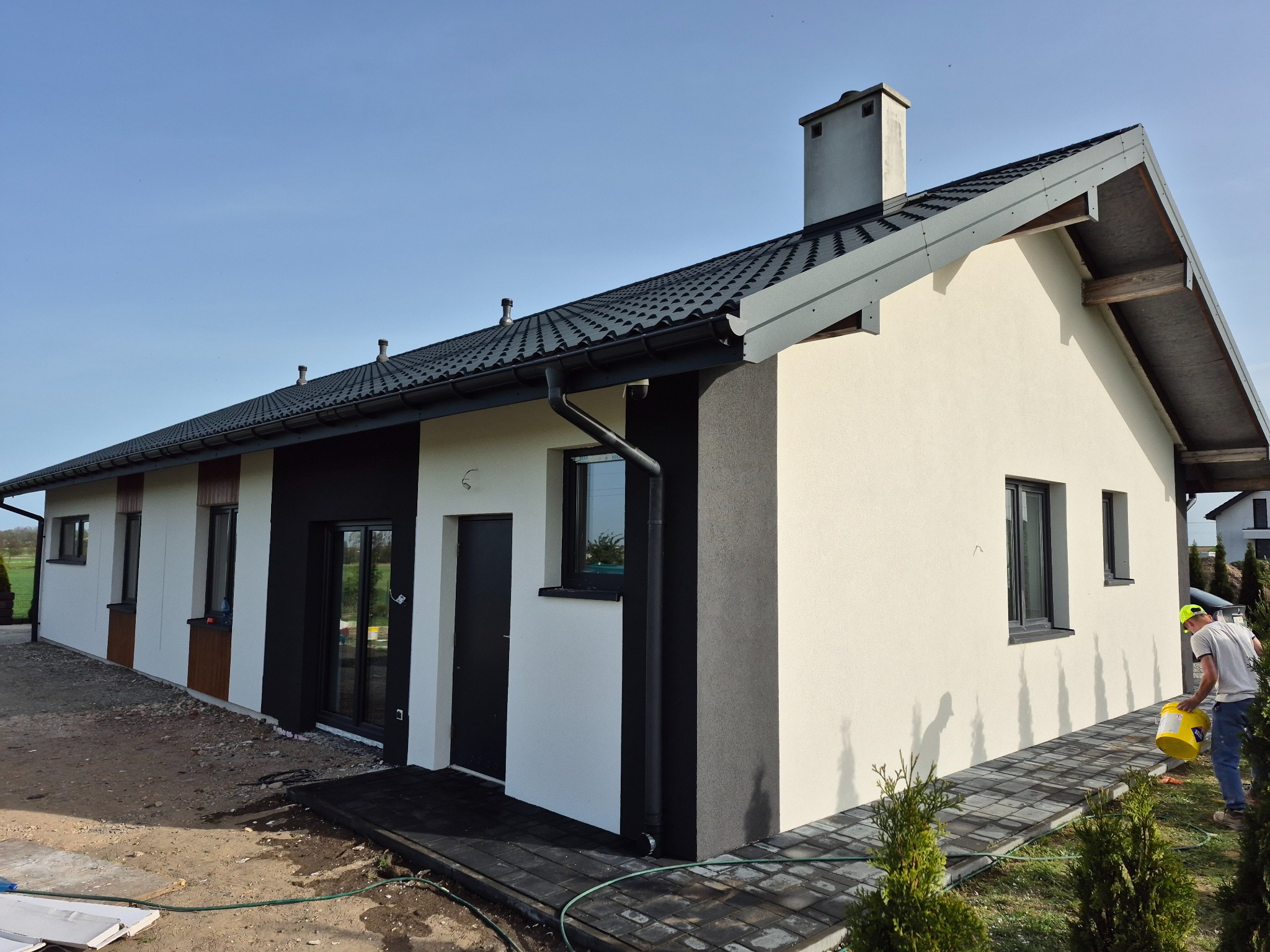 Jasny dom jednorodzinny z ciemną stolarką okienną i dachem. Osoba z żółtym wiadrem w tle. Elewacja w odcieniach bieli i szarości.
