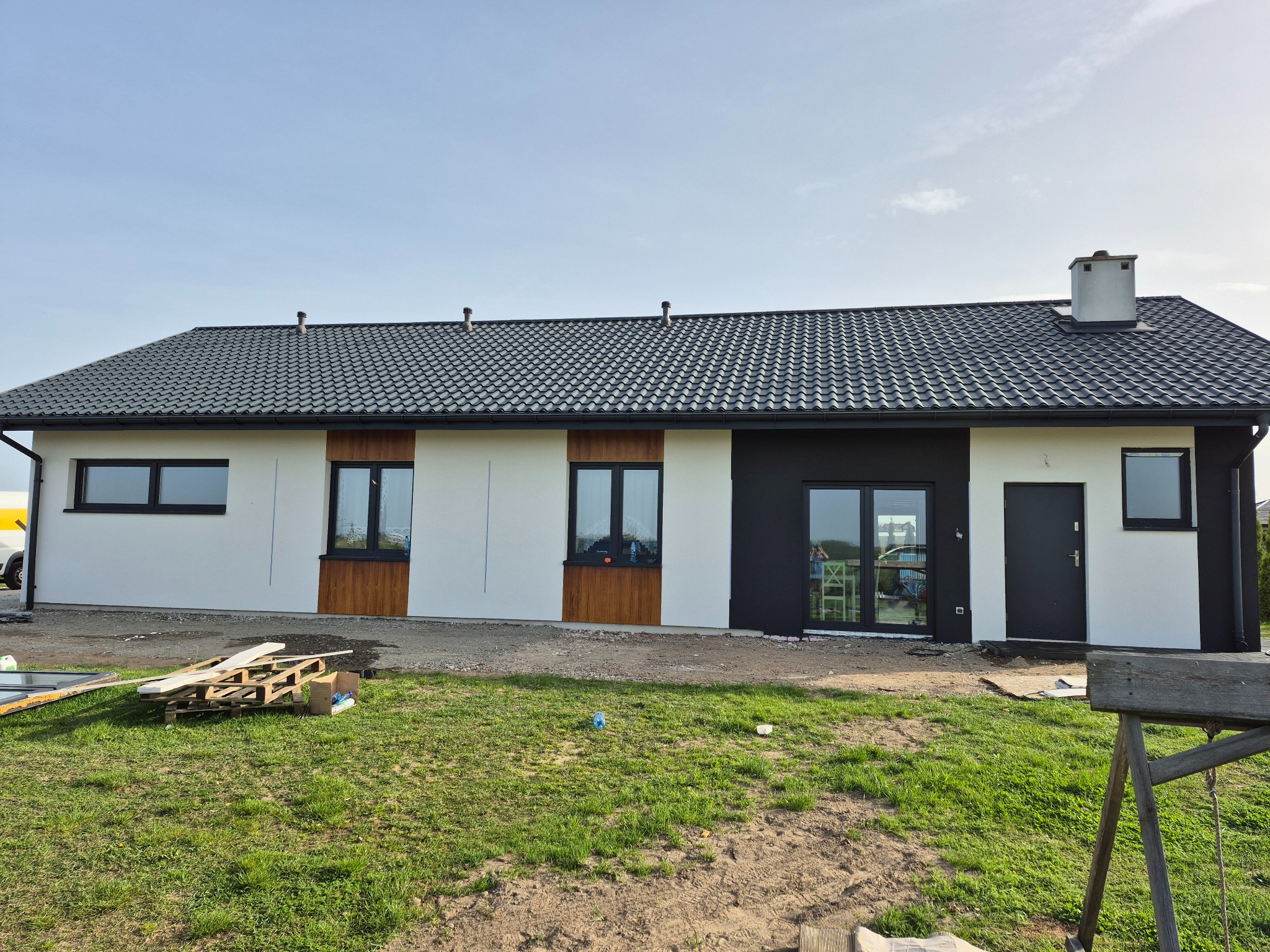 Nowoczesny dom jednorodzinny z białą elewacją, czarnymi detalami i grafitowym dachem. Widoczne okna z drewnopodobną okleiną i fragment placu budowy.