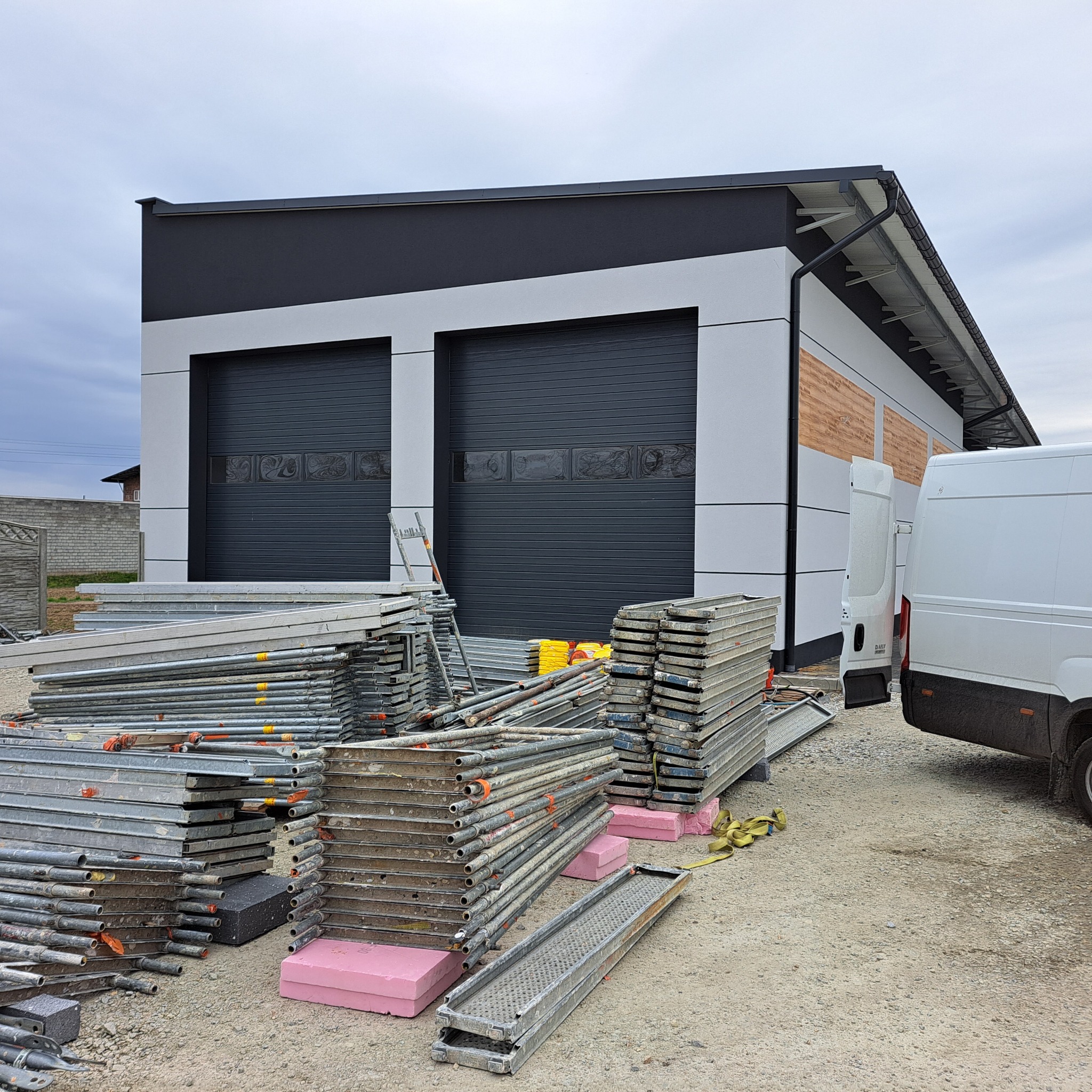 Budynek po wykonaniu elewacji w Młynisko. Widoczne rusztowania i samochód dostawczy. Szare panele elewacyjne i czarne bramy garażowe.