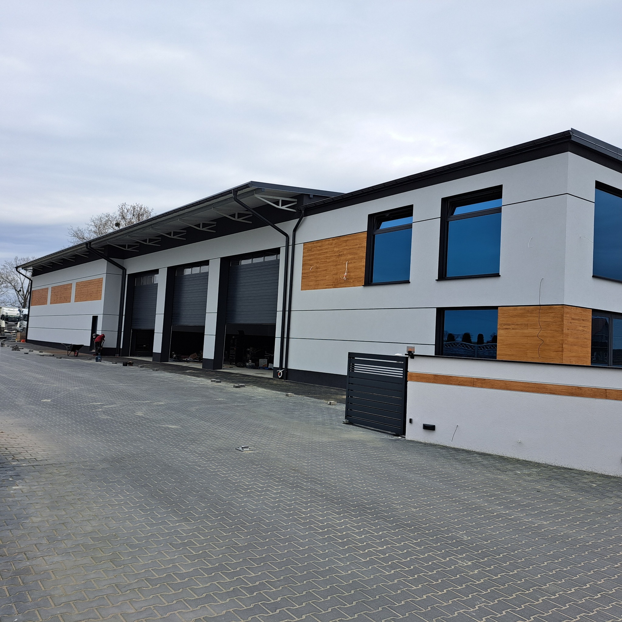 Nowoczesny budynek z szarą elewacją, drewnianymi akcentami i dużymi oknami. Widoczne bramy garażowe i kostka brukowa. Osoba pracuje na zewnątrz budynku.