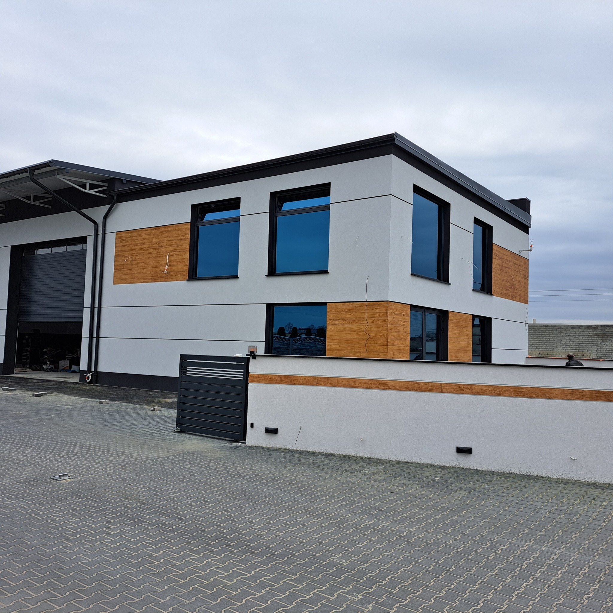 Nowoczesny budynek z elewacją w odcieniach szarości i drewnopodobnymi panelami, czarne ramy okien i brama wjazdowa. Widok z poziomu kostki brukowej.