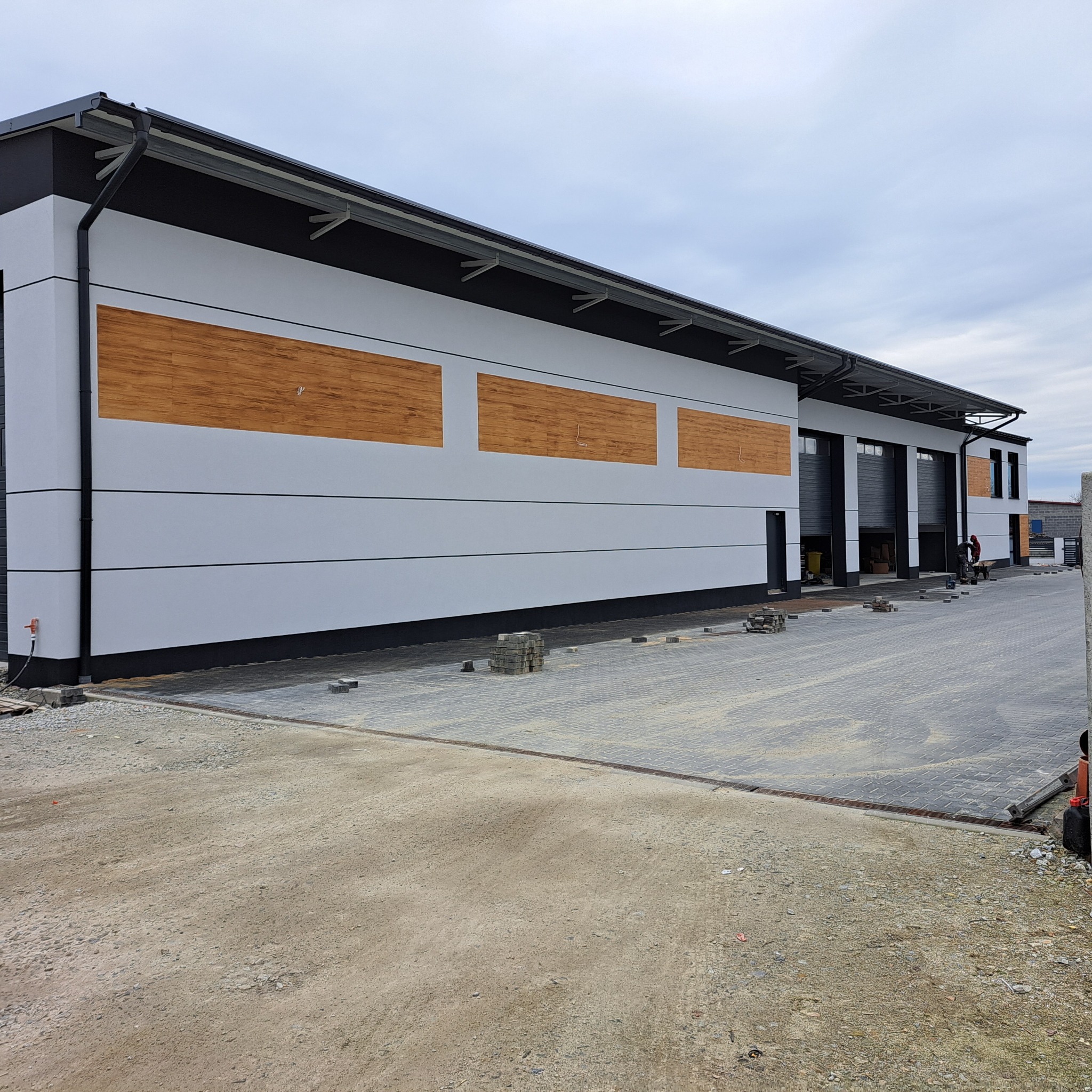 Nowoczesny budynek z elewacją w odcieniach bieli i czerni, z elementami dekoracyjnymi imitującymi drewno. Widoczne otwarte bramy garażowe i częściowo ułożona kostka brukowa.