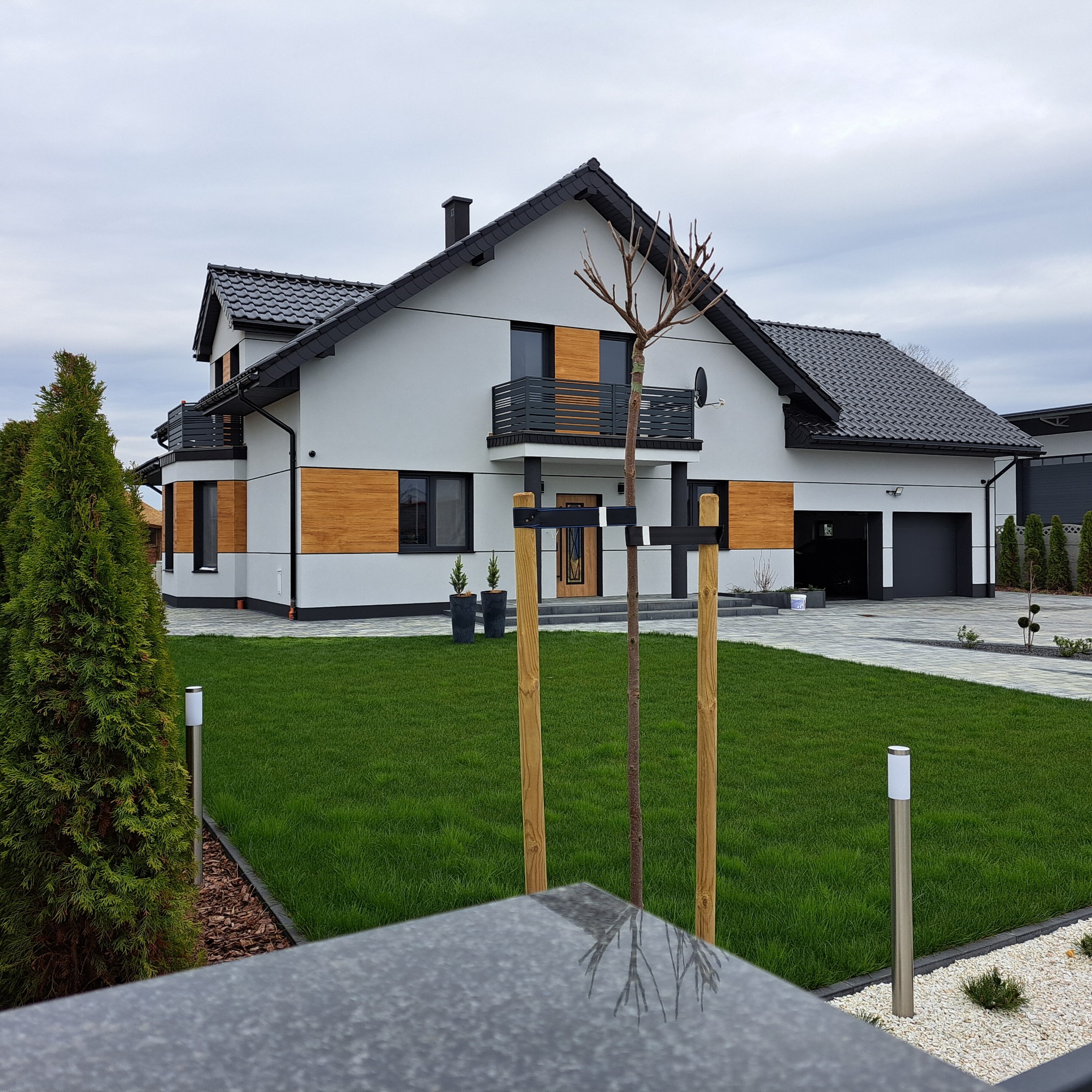 Nowoczesny dom jednorodzinny z grafitową dachówką i drewnianymi akcentami na elewacji, zadbany trawnik i oświetlenie zewnętrzne, widok z poziomu ogrodzenia.
