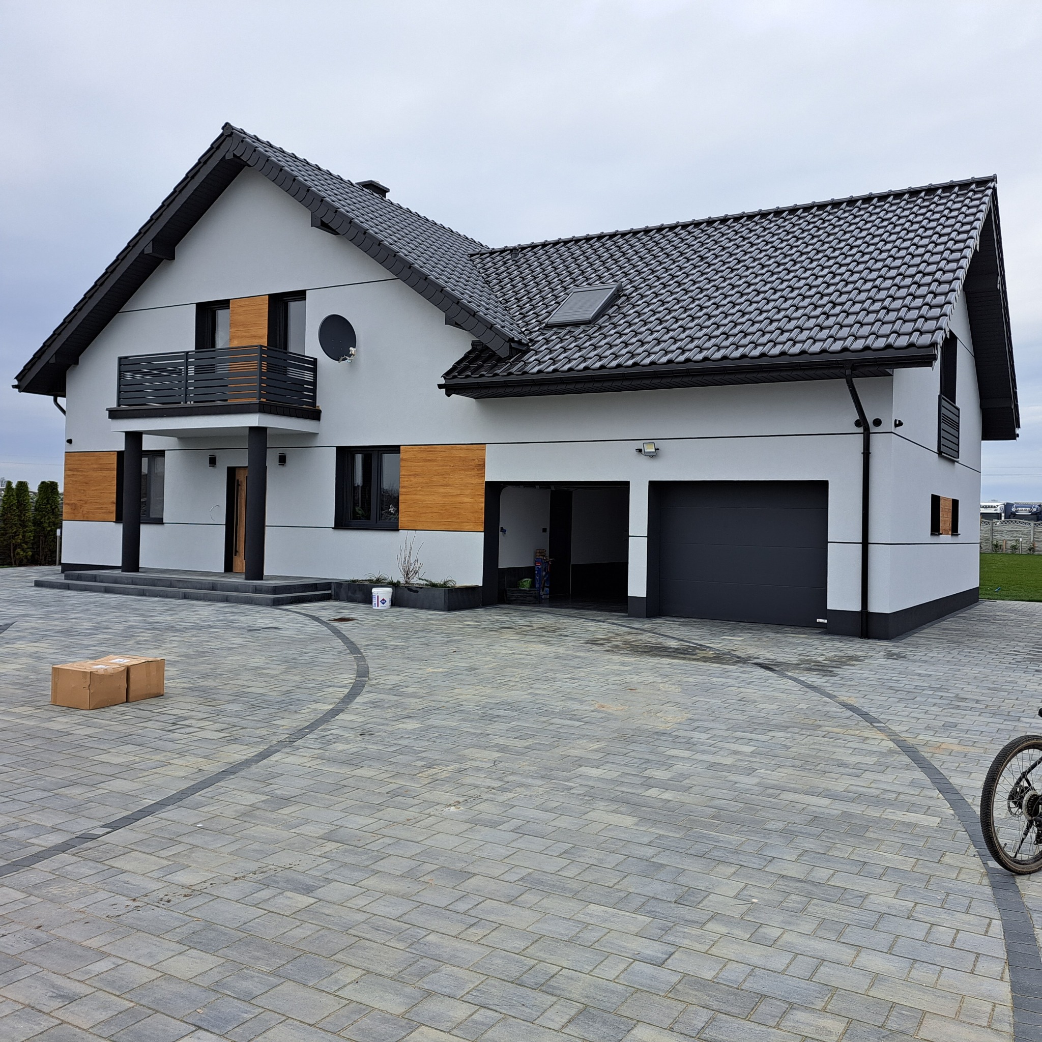 Nowoczesny dom z szarą elewacją i drewnianymi akcentami. Widoczny balkon, dachówka w kolorze antracytu i podjazd z kostki brukowej. Na podjeździe leżą kartony.