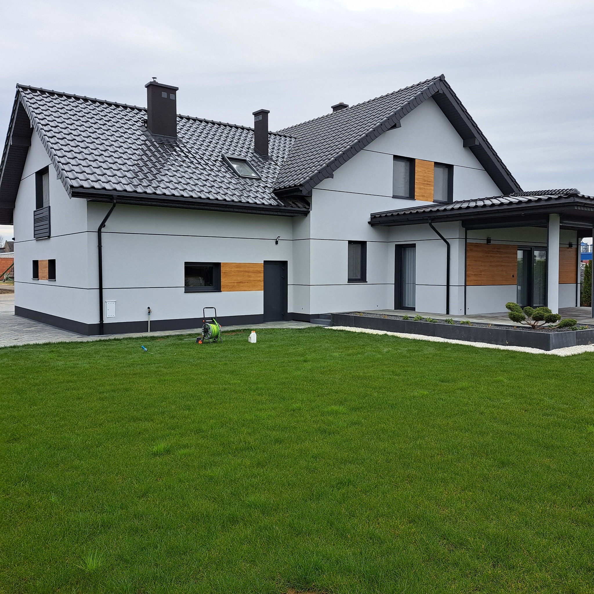 Nowoczesny dom z szarą elewacją, drewnianymi akcentami i ciemną dachówką, otoczony zielonym trawnikiem. W tle widoczne niebo i fragment sąsiedniego budynku.