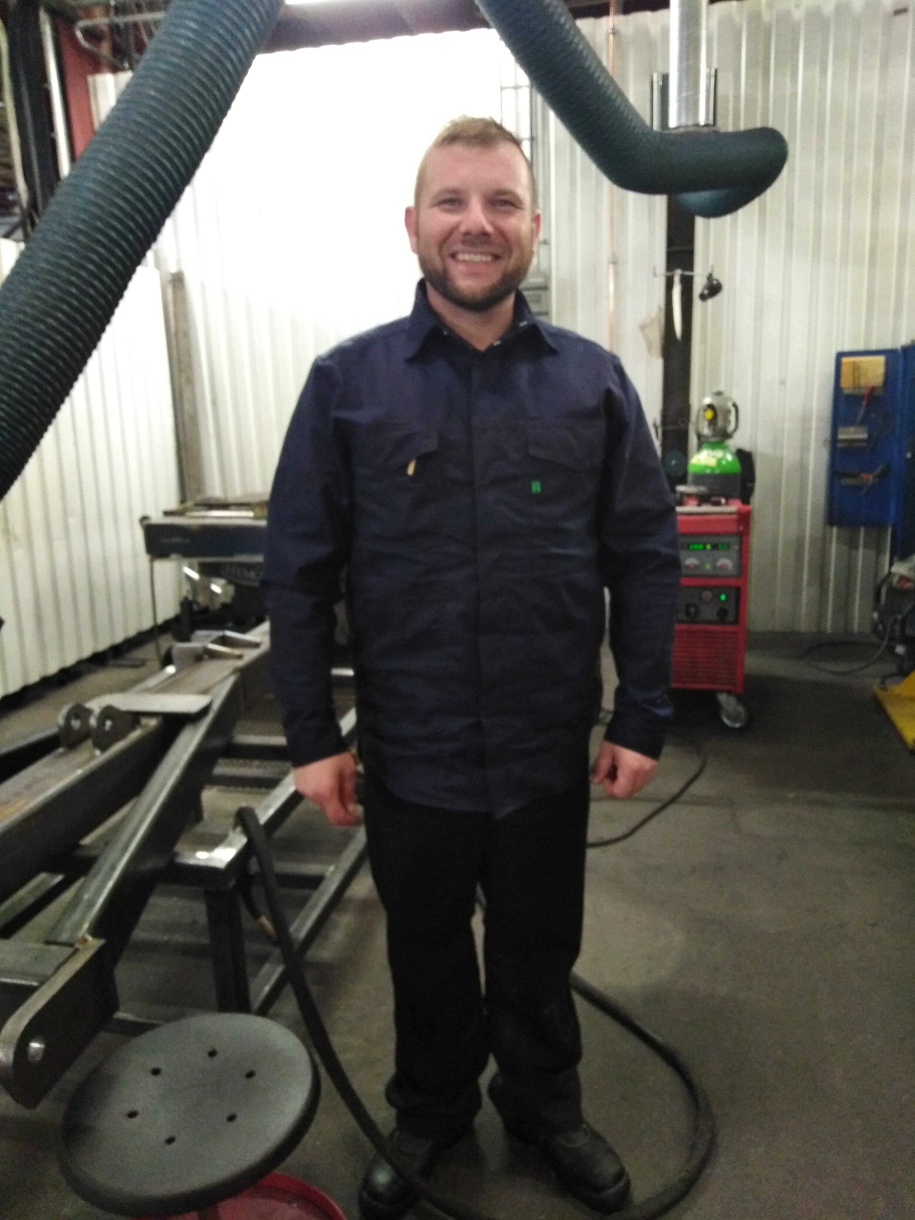 Uśmiechnięty mężczyzna w roboczym ubraniu na tle warsztatu z elementami konstrukcji metalowych i sprzętem spawalniczym.