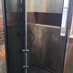 Nowoczesna kabina prysznicowa z czarnymi profilami i deszczownicą, wykończona płytkami imitującymi rdzę oraz mozaiką w kształcie plastra miodu.