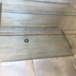 Zamknięta kabina prysznicowa z przesuwnymi drzwiami ze szkła hartowanego, zamontowana na jasnych, beżowych płytkach imitujących kamień. Widoczny odpływ liniowy w podłodze. U dołu kadru ceramiczna...