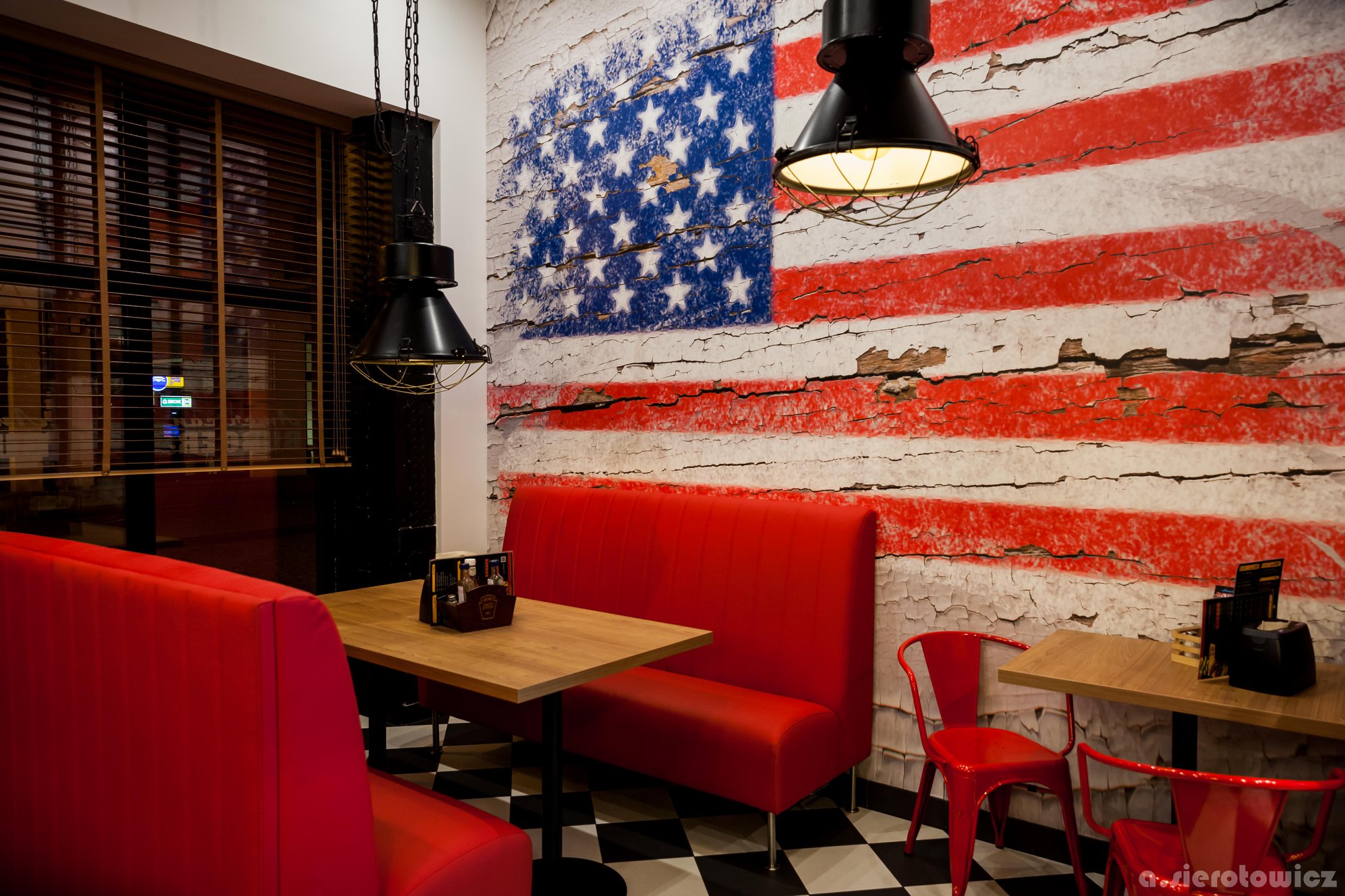 Wnętrze restauracji w stylu amerykańskim: czerwone kanapy, drewniane stoliki, czarno-biała podłoga w szachownicę i ściana z motywem flagi USA.