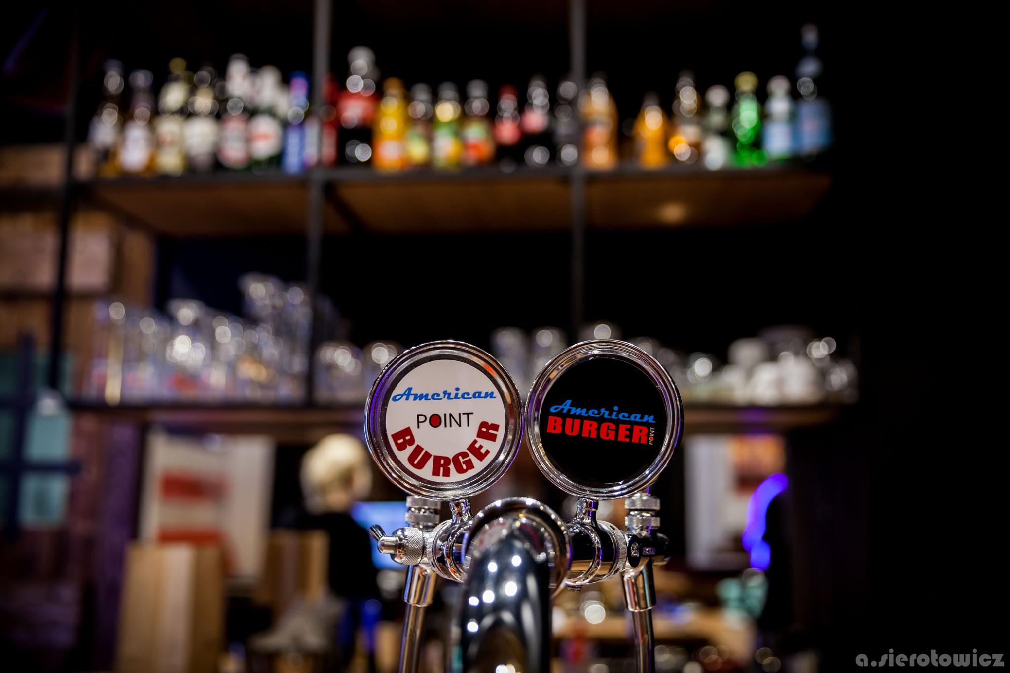 Dwa krany do piwa z logo American Point Burger na tle baru z półkami pełnymi butelek i szklanek.