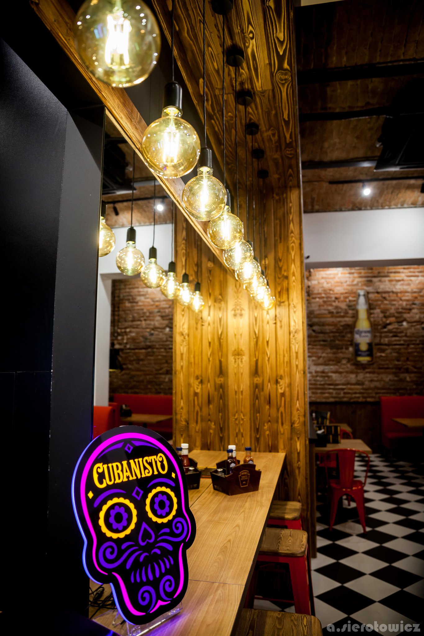 Wnętrze baru z drewnianym wystrojem, dekoracyjnymi żarówkami, neonowym znakiem 'Cubanisto' w kształcie czaszki i szachownicową podłogą.