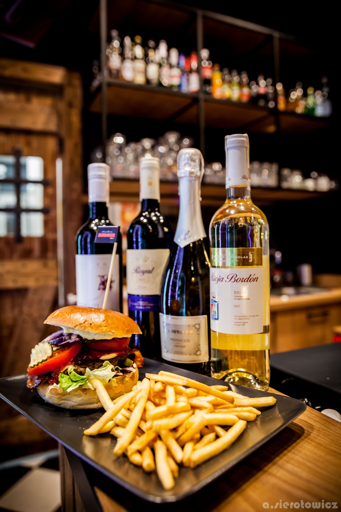 Burger z frytkami na czarnym talerzu, w tle butelki wina i szampana, drewniane wykończenie baru i półki z alkoholem.