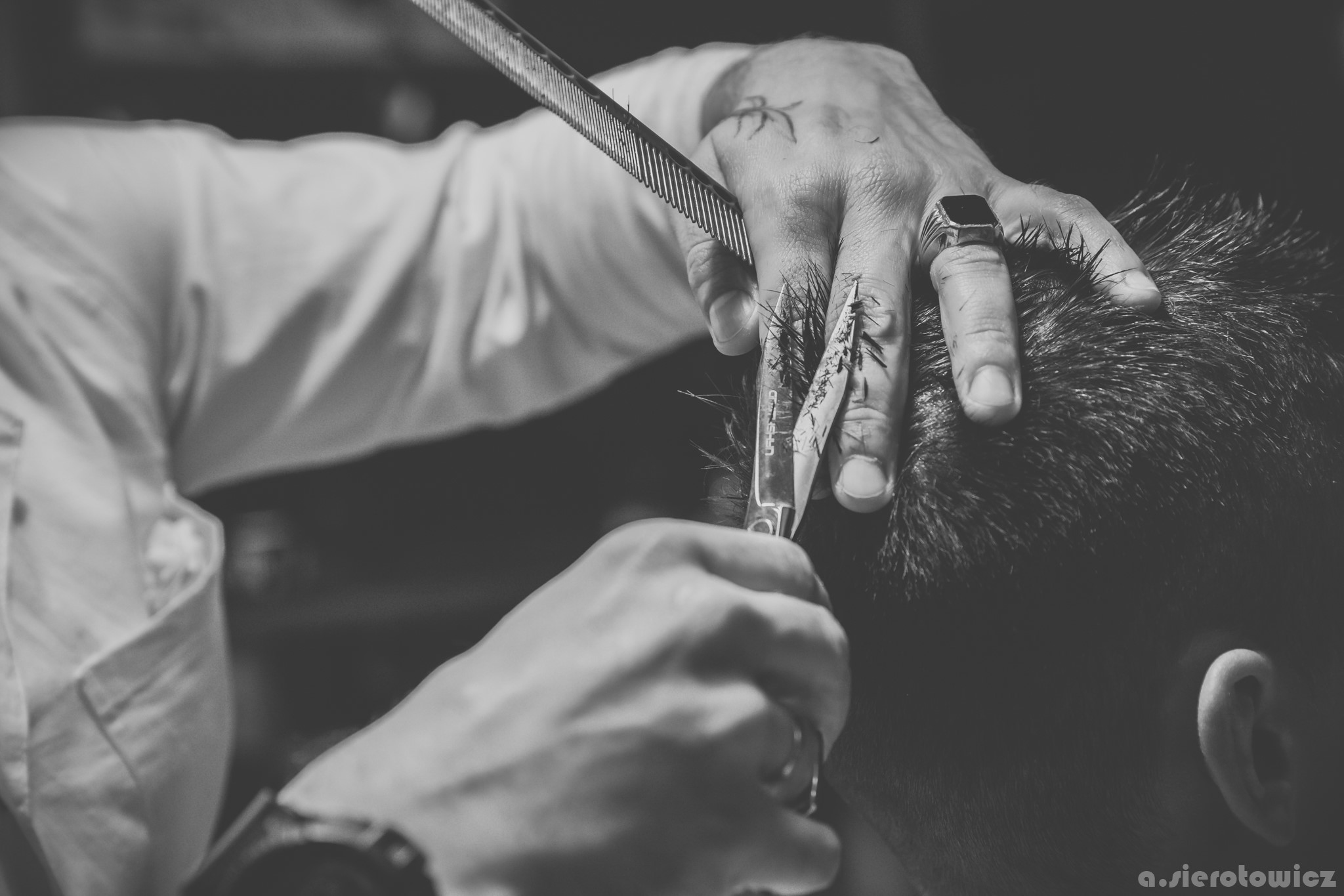 Czarno-białe zdjęcie fryzjera w trakcie strzyżenia włosów klientowi, widoczne ręce z tatuażami, grzebień i nożyczki, zbliżenie na włosy.