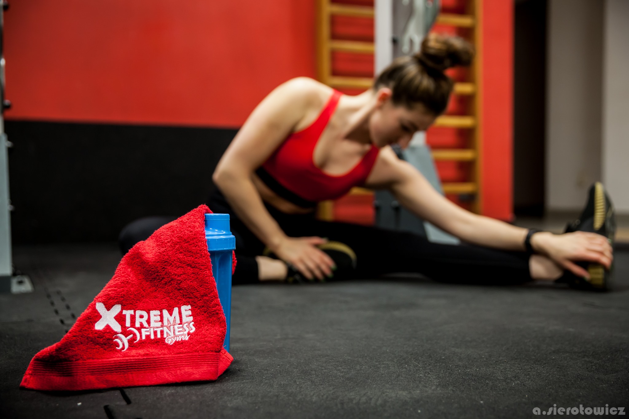 Kobieta w czerwonym sportowym topie rozciąga się na podłodze w siłowni, w tle drabinki gimnastyczne, na pierwszym planie czerwony ręcznik z logo 'Xtreme Fitness Gyms' i niebieski shaker.