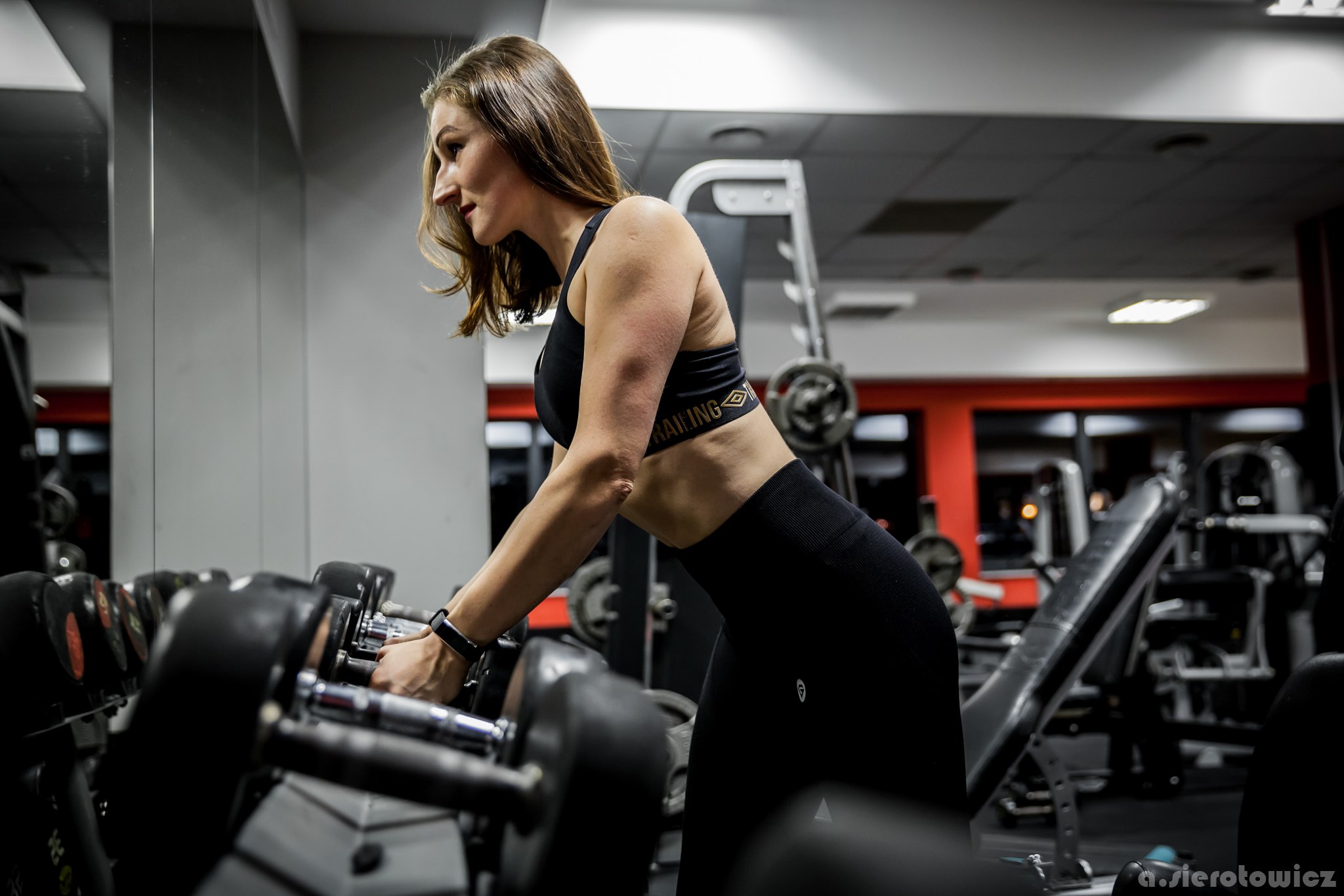 Kobieta w sportowym stroju, pochylona nad rzędem hantli w siłowni, z widocznym zegarkiem sportowym na nadgarstku.