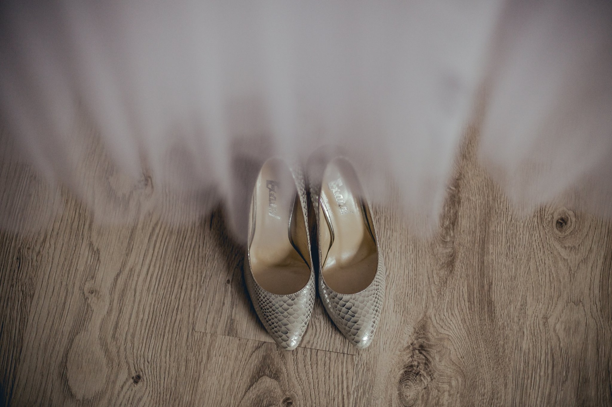 Eleganckie, srebrne szpilki ze wzorem skóry węża na drewnianej podłodze, częściowo zasłonięte przez miękki, rozmyty tiul.