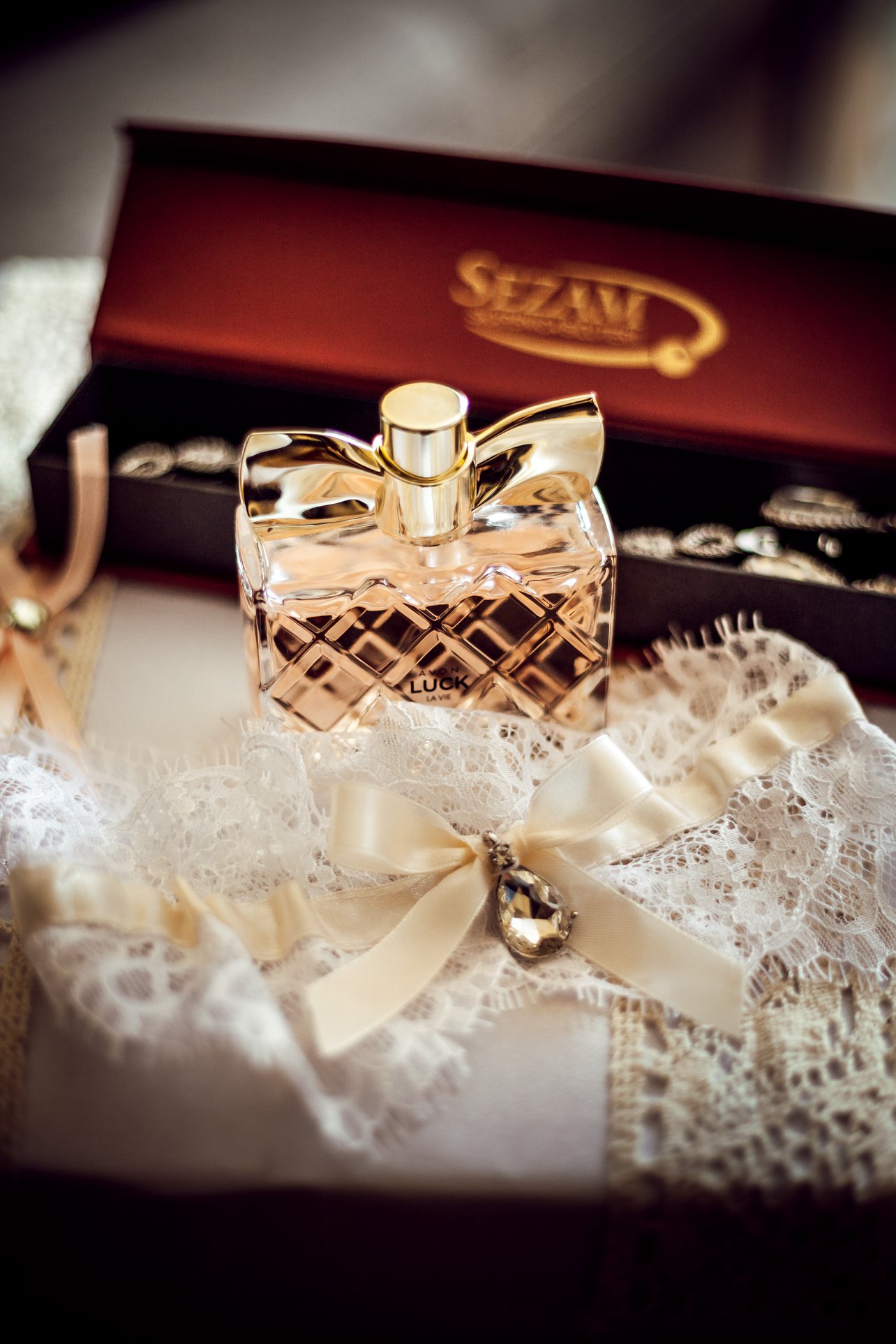 Perfumy z kokardą na koronkowej podwiązce z kryształkiem, w tle pudełko na biżuterię.