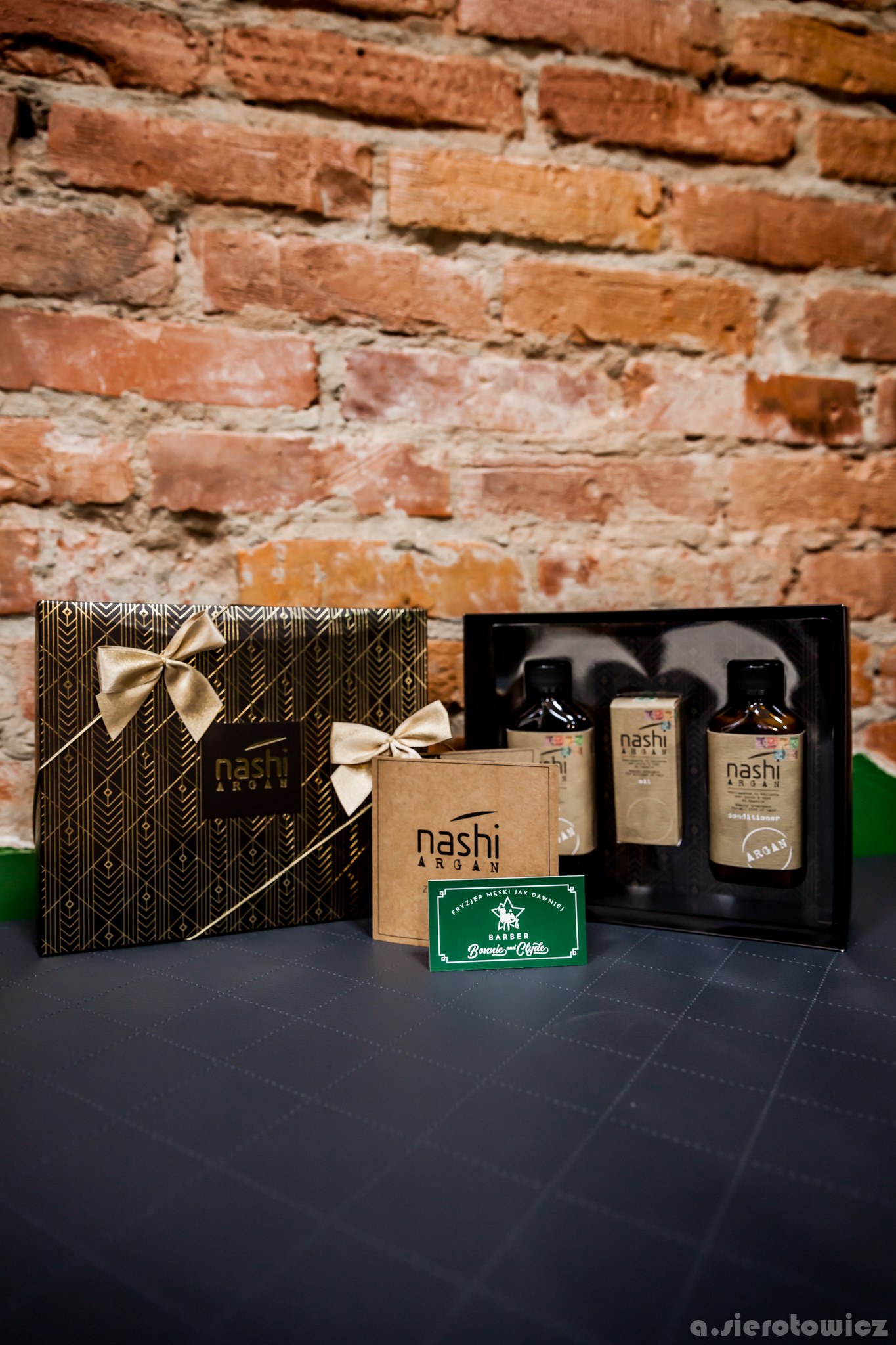 Zestaw kosmetyków Nashi Argan w eleganckich opakowaniach na tle ceglanej ściany, z wizytówką salonu fryzjerskiego Bonnie & Clyde.