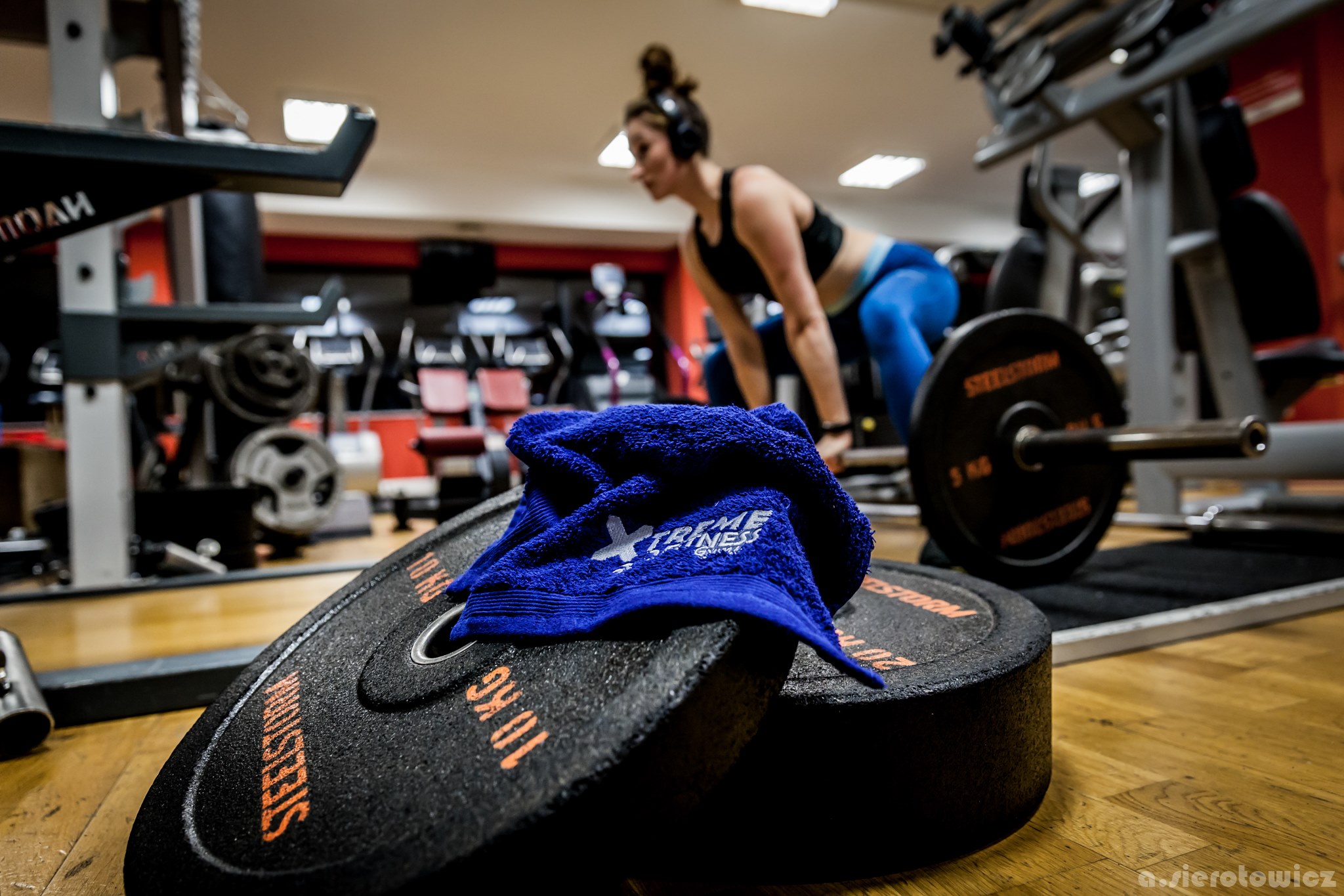 Dwa czarne obciążenia 10 kg z niebieskim ręcznikiem Xtreme Fitness na podłodze siłowni, w tle kobieta w słuchawkach przygotowująca się do podnoszenia ciężarów.