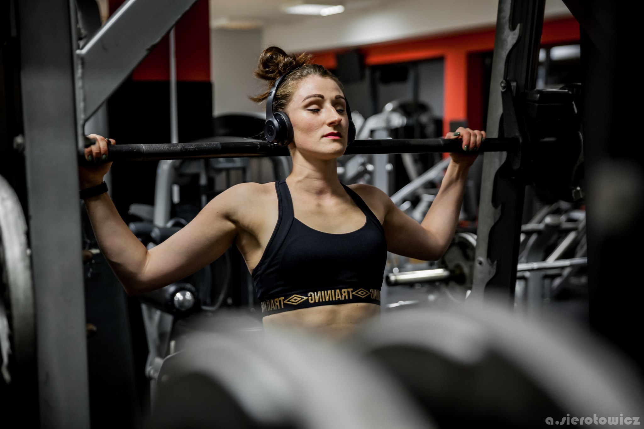Kobieta w czarnym sportowym topie i słuchawkach, podnosi sztangę na siłowni, skupiona z zamkniętymi oczami.