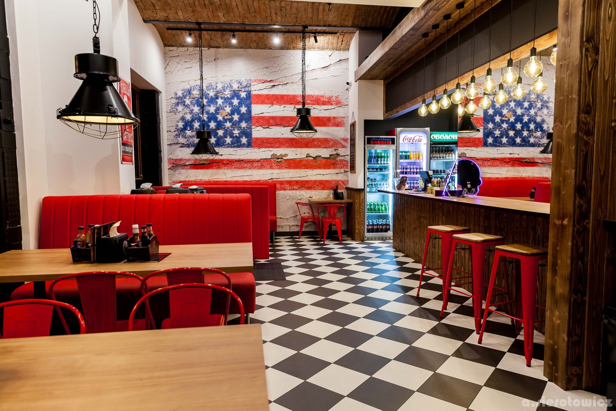 Wnętrze restauracji w stylu amerykańskim z czerwonymi meblami, ścianą z motywem flagi USA i czarno-białą podłogą w szachownicę, oświetlone wiszącymi lampami.