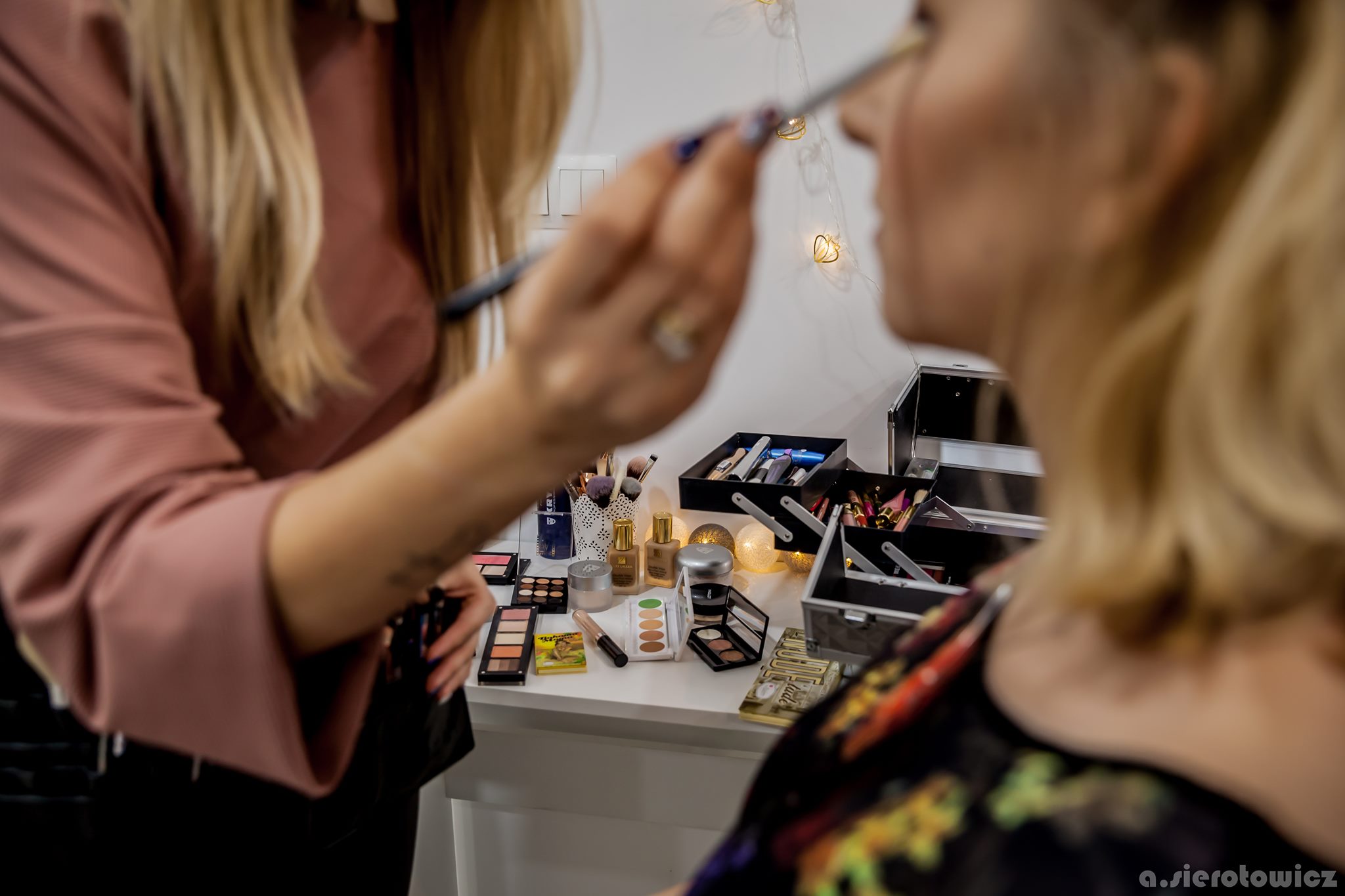 Wizażystka z pierścionkiem na palcu, w różowej bluzce, aplikuje pędzlem makijaż na powiekę klientki, na białym stole rozłożone kosmetyki i pędzle, w tle lampki.