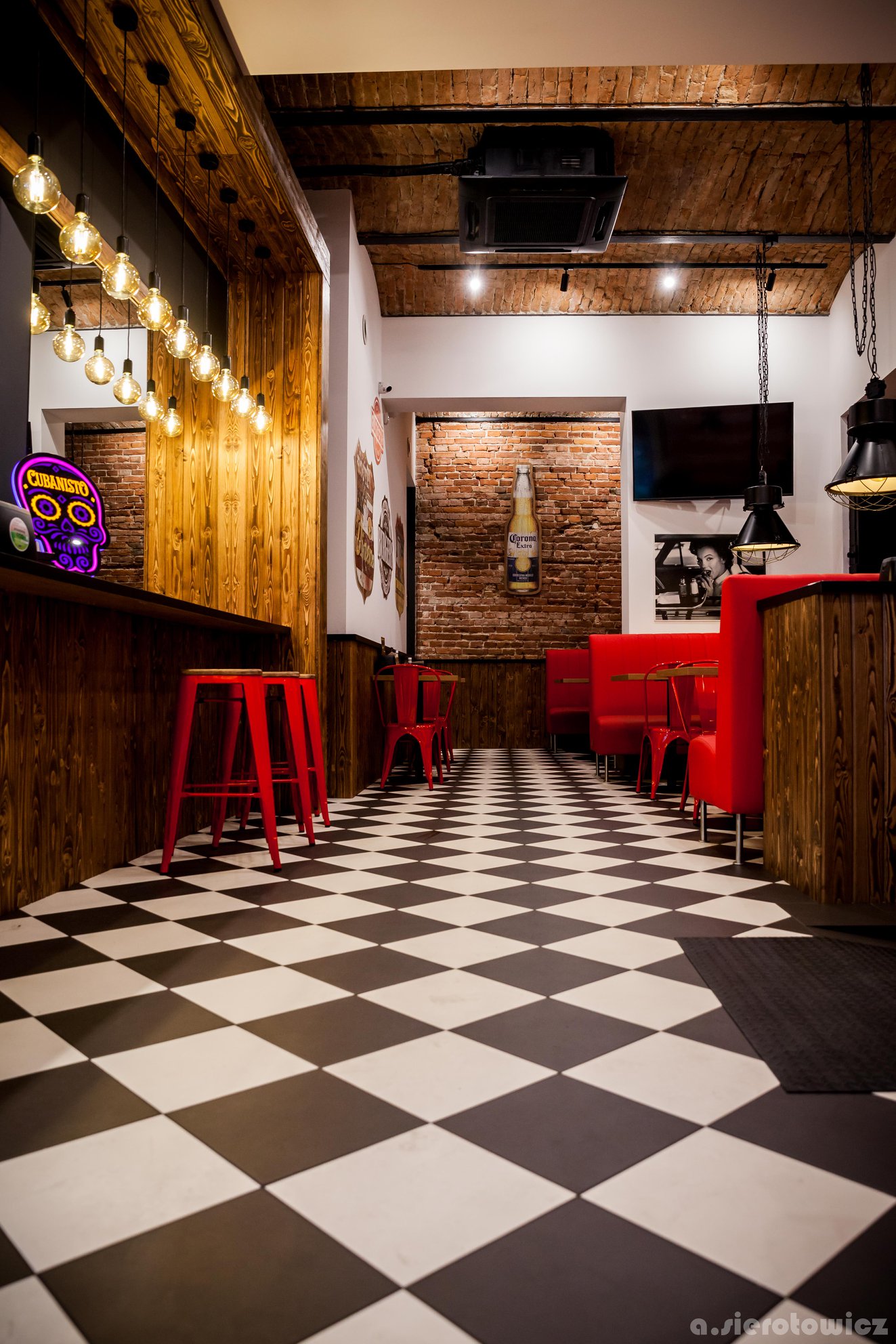 Wnętrze lokalu gastronomicznego z czarno-białą szachownicą na podłodze, czerwonymi krzesłami i lożami, drewnianymi elementami wystroju, ceglanymi ścianami i dekoracyjnym oświetleniem, prezentujące...