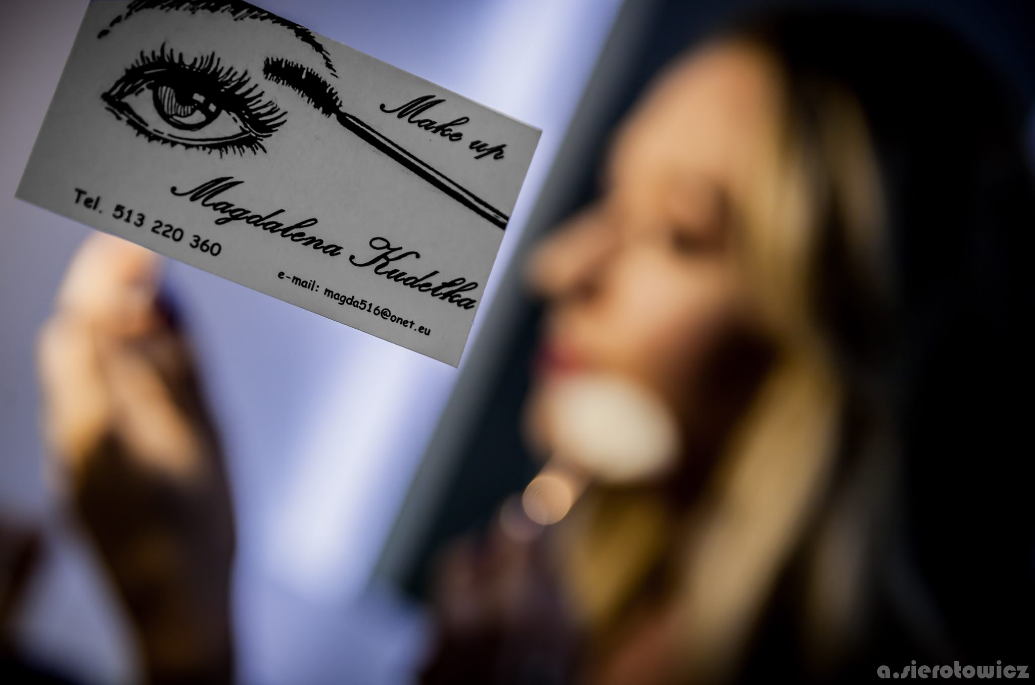 Wizytówka makijażystki z ilustracją oka i tuszu do rzęs, trzymana na tle rozmytej twarzy kobiety podczas wykonywania makijażu.