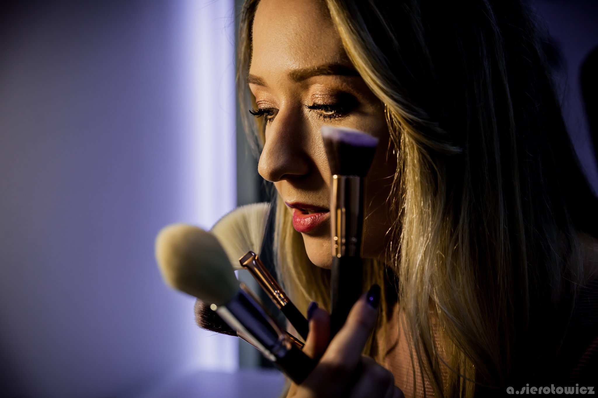 Profil kobiety z blond włosami, trzymającej zestaw pędzli do makijażu w dłoni, na tle fioletowej ściany.